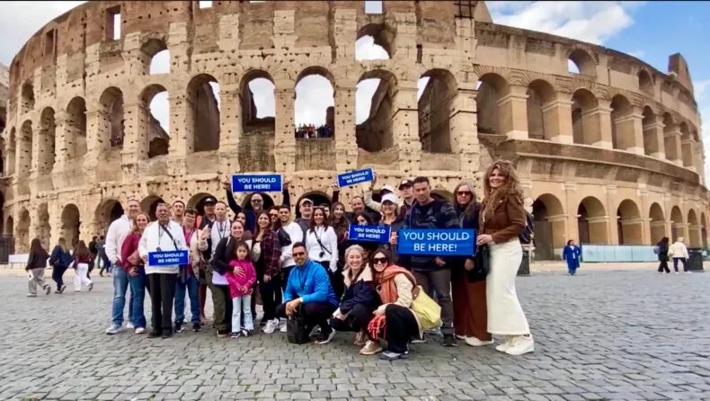 DreamTrips travelers posing in front of the Colosseum in Rome holding You Should Be Here signs