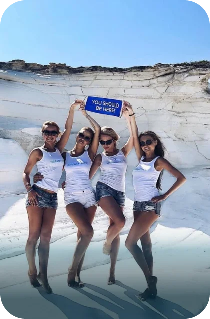 Four women holding the iconic “You Should Be Here” sign during a DreamTrips experience at beach cliffs.