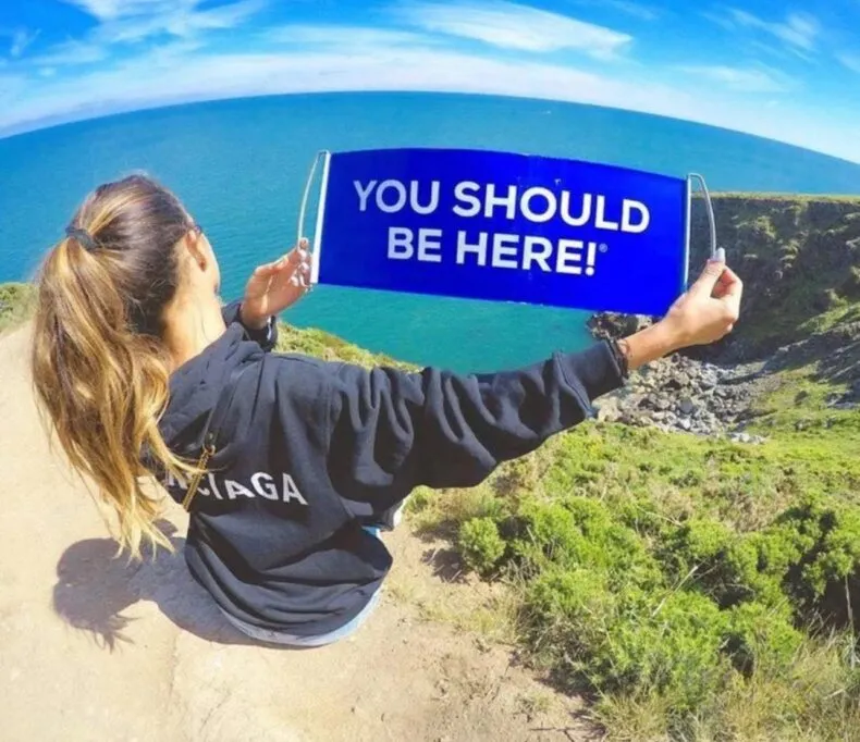Traveler holding a blue “You Should Be Here” sign overlooking a scenic ocean cliff