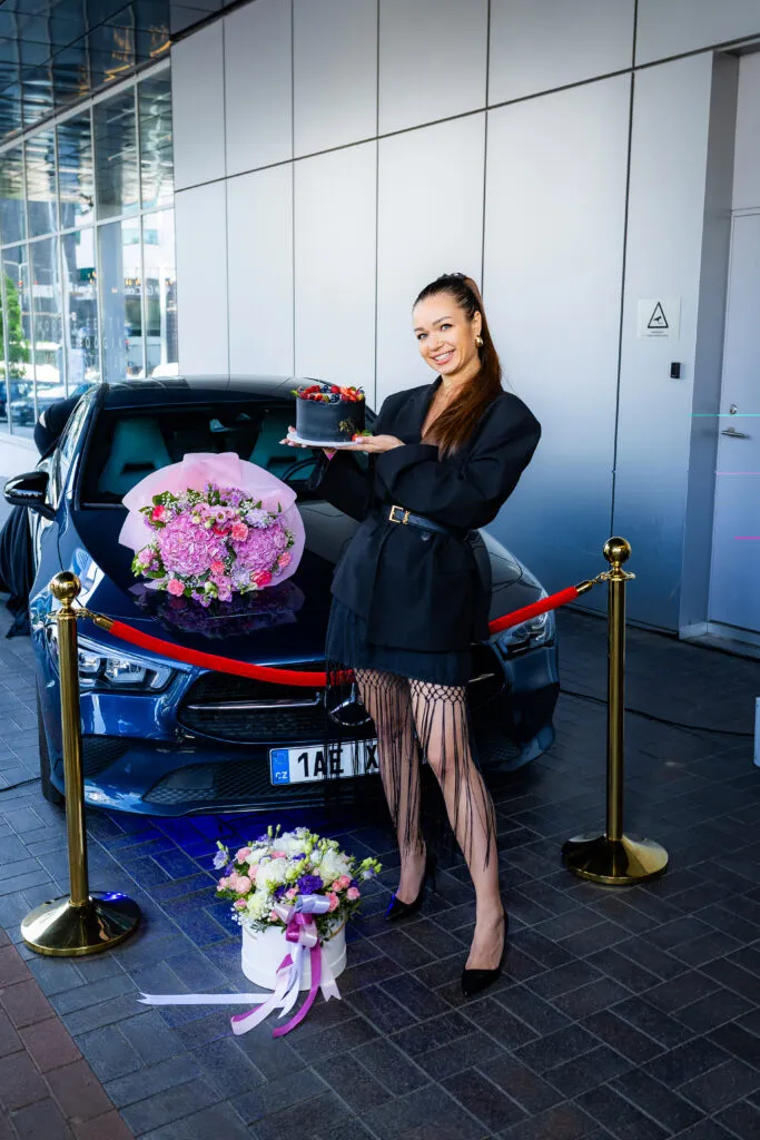 Greenway Global leader holding a cake in front of a car, surrounded by flowers and red ribbon at a recognition ceremony.