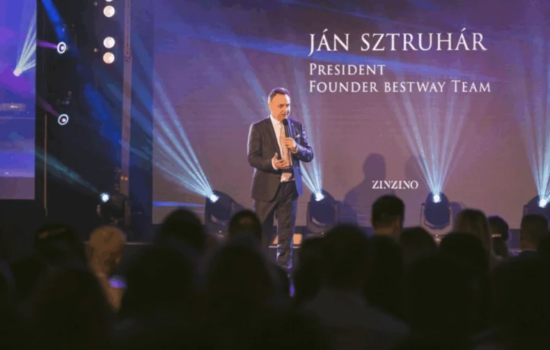János Sztruhár speaking on stage during a Zinzino event, holding a microphone with blue lights and audience in view.