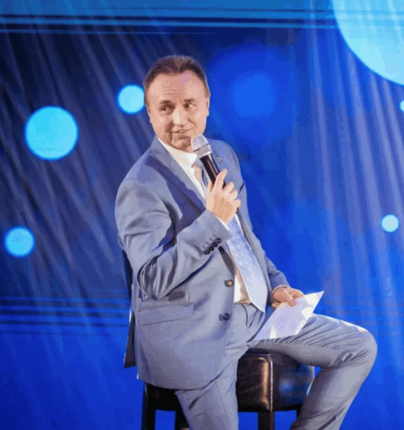 János Sztruhár sitting on stage with a microphone, wearing a blue suit during a Zinzino event.