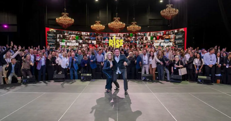Steve and Gina Merritt on stage with a large LifeWave team celebrating at a leadership event, with “Rise” and “One Million” signs in the background.